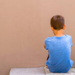 Sad alone boy sitting on the ground behind the wall outdoor.